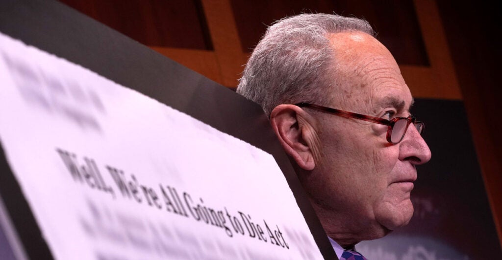 Chuck Schumer in profile next to a display reading "Well, We're All Going to Die Act"