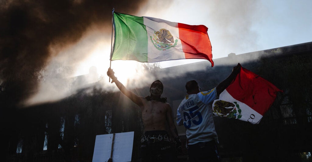LA Riot. As smoke rises behind them protesters wave Mexican flags.