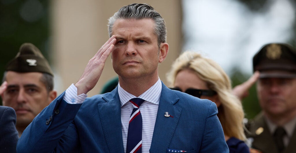 Pete Hegseth in a blue suit salutes at D-Day ceremonies.