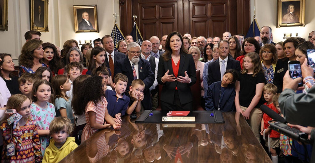 New Hampshire Gov. Kelly Ayotte, surrounded by children, stands at a desk.