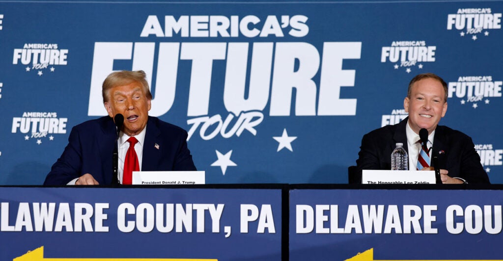 Donald Trump and Lee Zeldin sit together on a dias at a 2024 campaign event in front of sign reading "America's Future Tour."