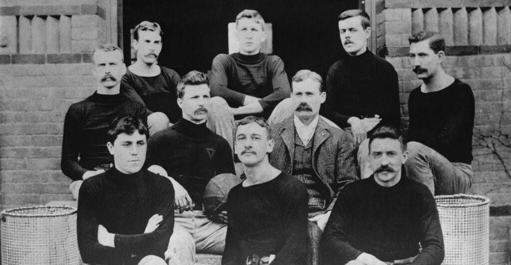 A 1891 shot of James Naismith in a suit sitting on steps with the first basketball team. The young men decked in black long sleeve shirts and tan trousers.