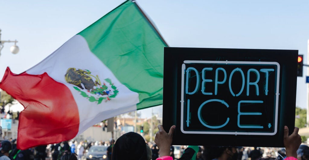Protester holds up a "Deport ICE" sign as a Mexican flag waves in the background.