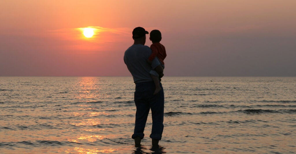 A silhouette of a father holding his son at the shoreline watching a sunset.