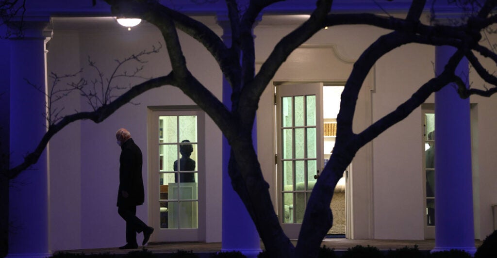 President Joe Biden in silhouette leaves Oval Office at night, as lights remain on and woman stands, presumably still working.