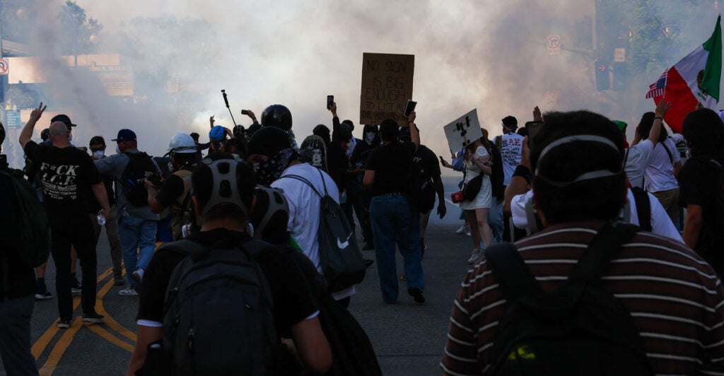 Shot from behind, a swarm of anti-ICE, anti-Trump protesters moves toward police in a street filled with smoke.