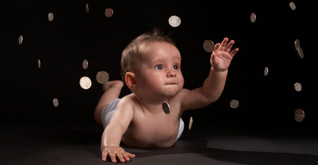Infant boy on his belly against a black background reaches in the air as coins fall around him.