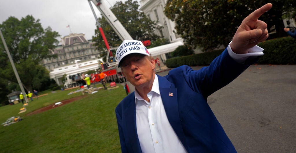 Donald Trump in a white "Make America Great Hat" and open collar shirt points as work is underway to put up a flagpole on the White House lawn.