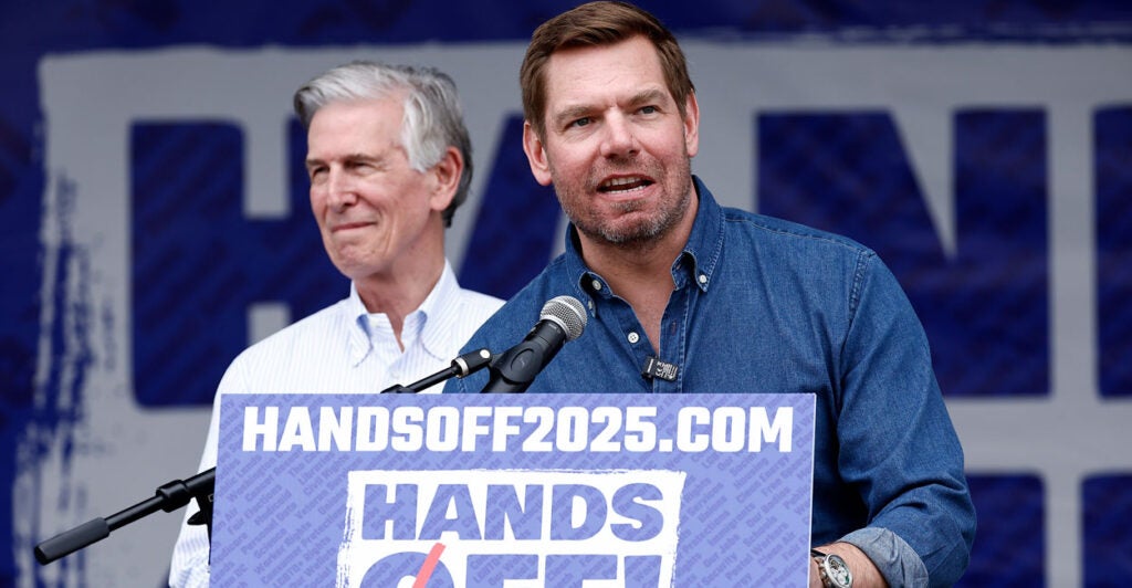 Eric Swalwell speaks behind a podium reading "Hands Off 2025.com" A white haired man stands behind him.