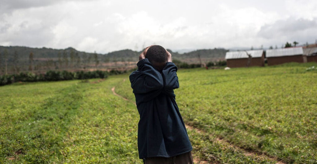 A Nigerian man in a field holds the back of his head with both hands, arms covering his face in grief.