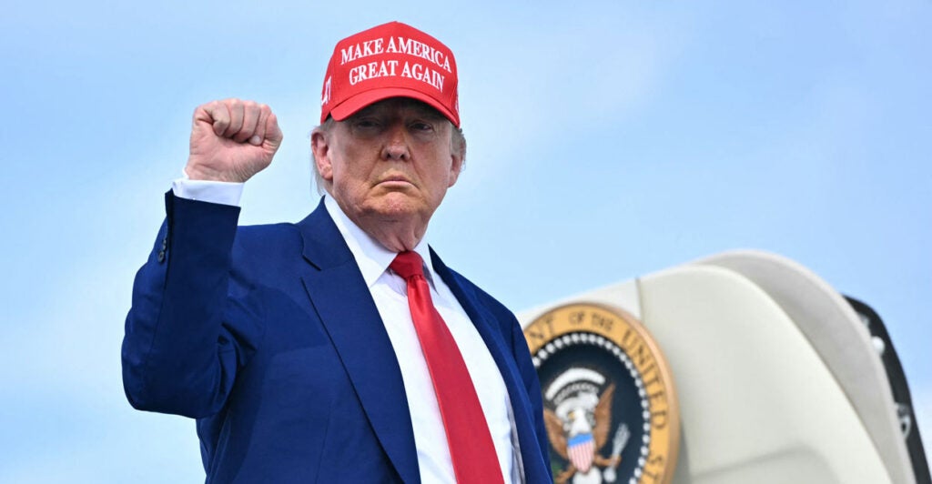 President Trump raises his fist while boarding Air Force One and waring a MAGA hat, red tie, and dark blue suit.