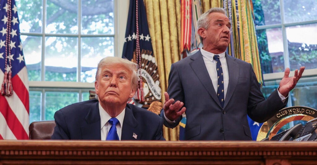 President Donald Trump gazes up while sitting at the Resolute Desk in Oval Office. Standing to his left and looking left is Robert F. Kennedy Jr., with arms wide.