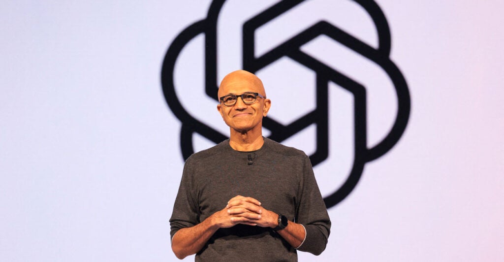 Microsoft CEO Satya Nadella in a gray shirt stands with his hands together in front of the OpenAI logo.