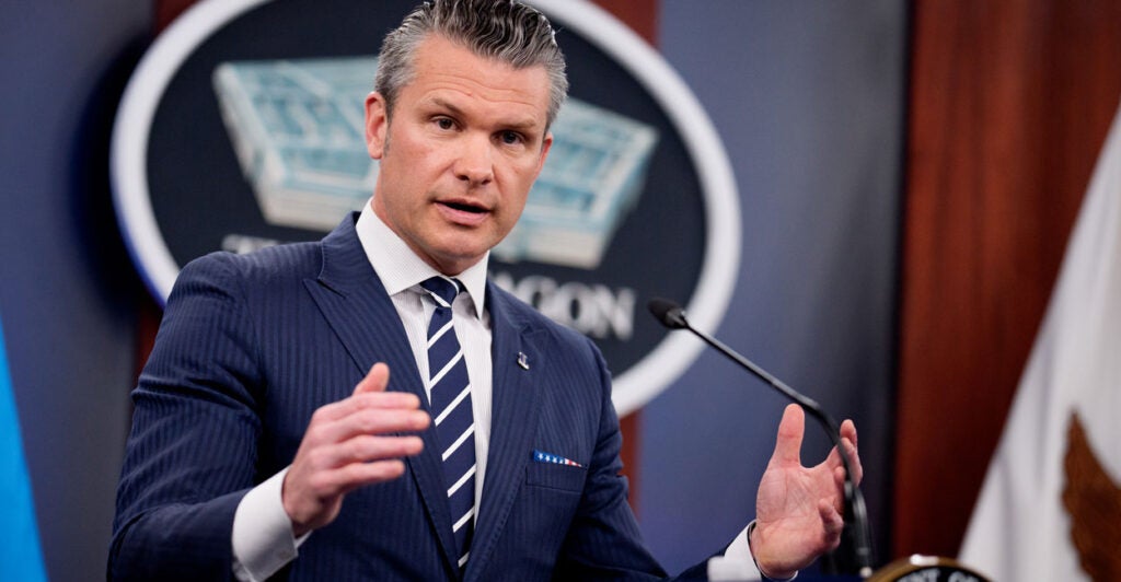 Defense Secretary Pete Hegseth in a blue suit speaks, with arms wide, at a Pentagon press briefing.