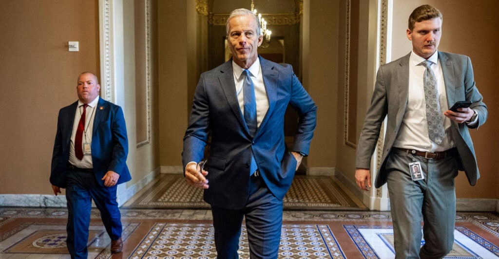 Senate Majority Leader John Thune strolls down a Senate corridor, flanked by two other people.