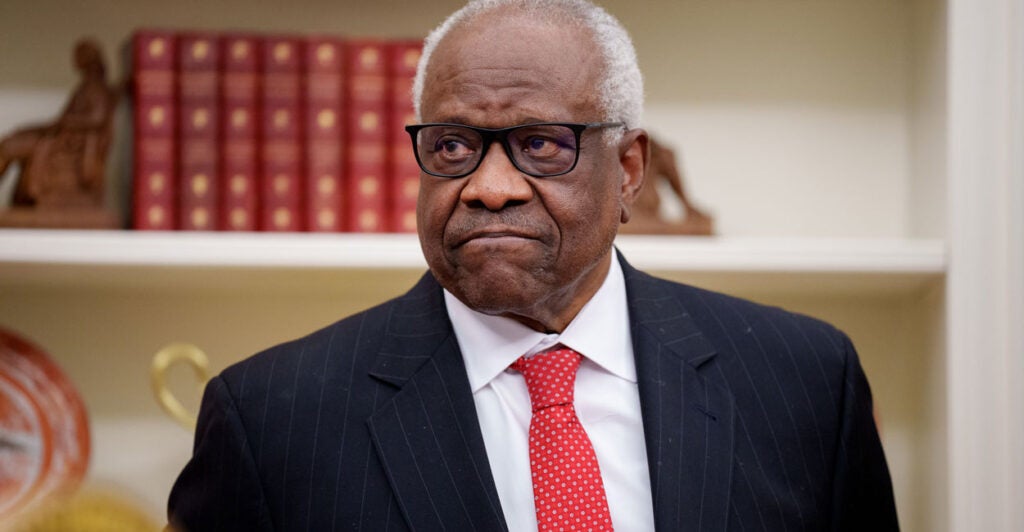 Supreme Court Justice Clarence Thomas, wearing a red tie, with what appear to be law books on a shelf behind him