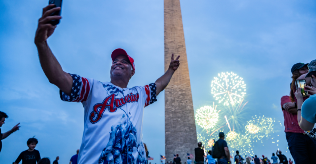 A man in an "America" jersey takes a selfie in front of the Washington Monument with fireworks and a crowd in the background.