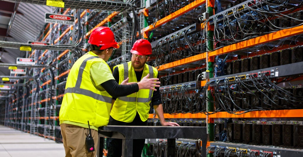 two employees in yellow fluorescent vests and red hardhats have a discussion inside a data center with thousands of computer servers, wires, and "danger -high voltage" signs