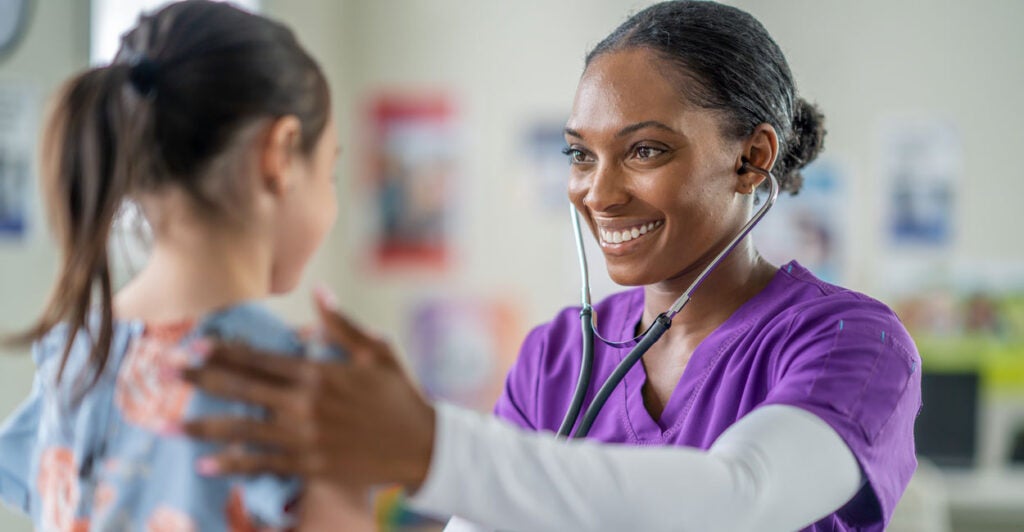 a black nurse in purple scrubs listens to the heartbeat of a young girl with a stethoscope