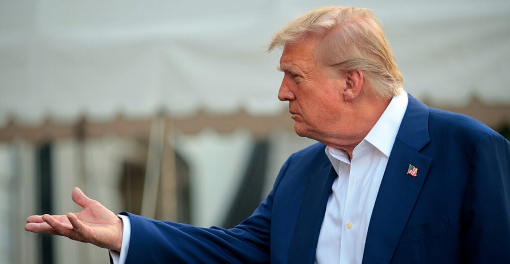 A side shot of President Trump in a blue suit with open white shirt holding out his hand.