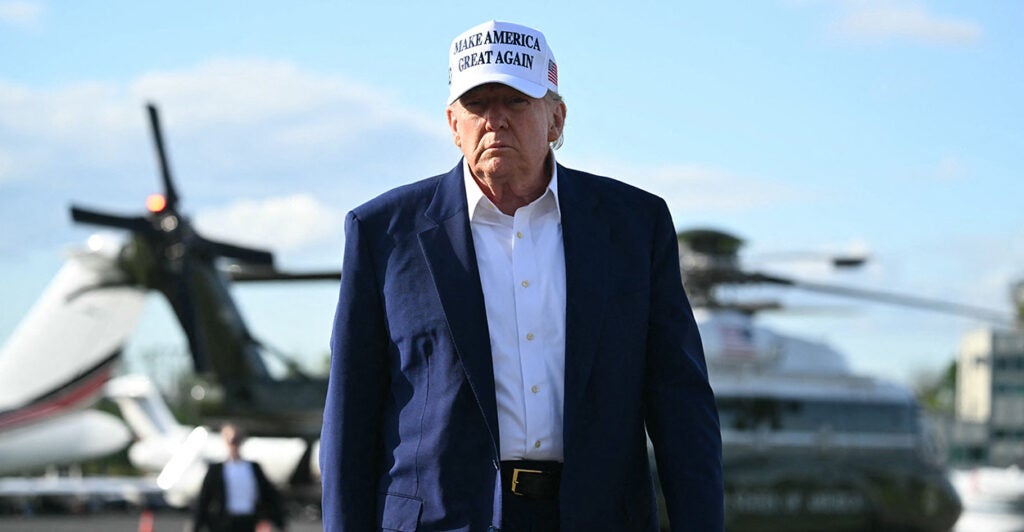 President Donald Trump in blue jacket, white shirt and white MAGA hat walks toward camera, with Marine One behind him.