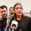 Alexandria Ocasio-Cortez stands at a podium in front of a microphone and looks to her left as she speaks.