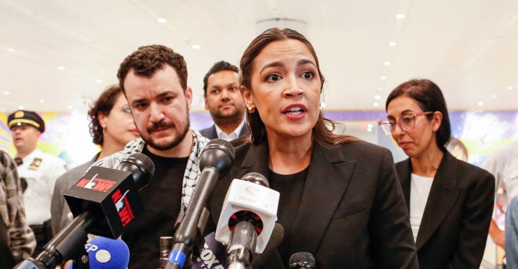 Alexandria Ocasio-Cortez stands at a podium in front of a microphone and looks to her left as she speaks.