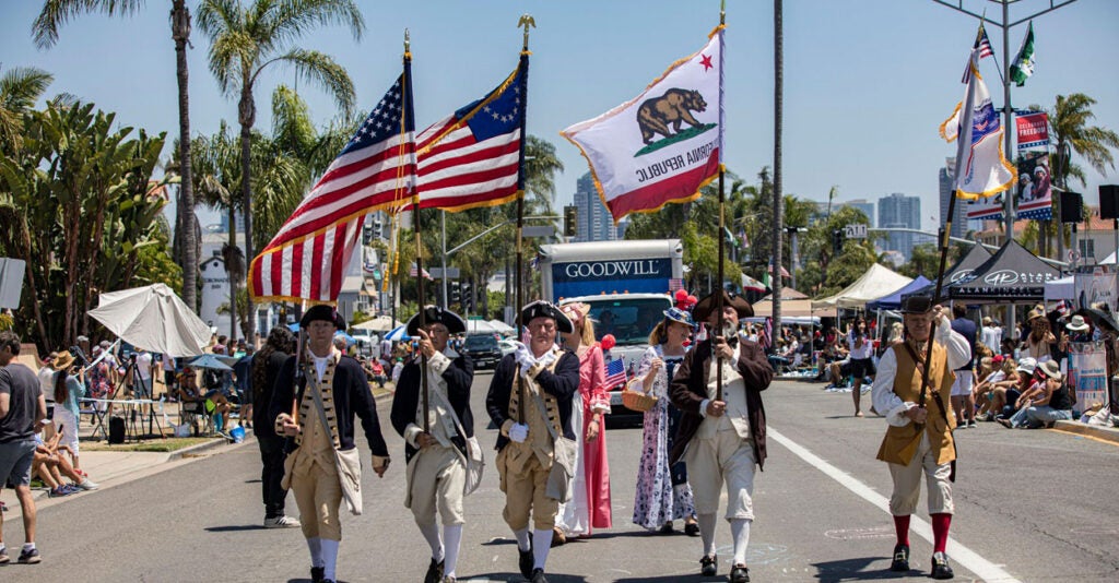 A group of men dressed like Minute Men from the Revolutionary War march down the street in a parade holding the American flag.