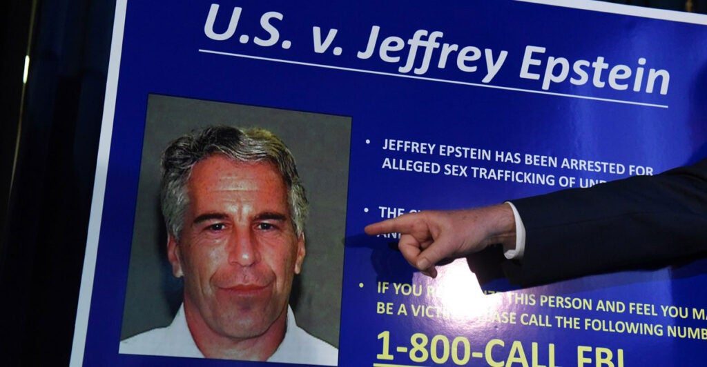 A finger points to a mugshot photo of Jeffrey Epstein on a poster board.