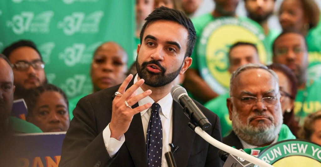 Zohran Mamdani stands and talks into a microphone and gestures with his right hand.