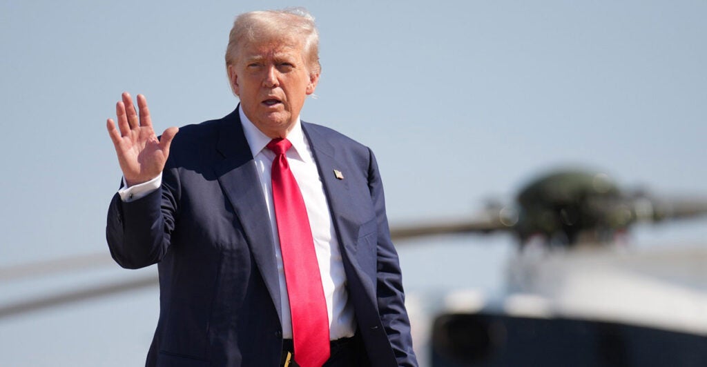 Donald Trump stands and holds his right hand out while a helicopter is in the background.