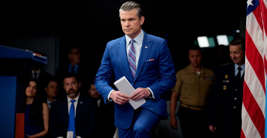 Pete Hegseth, in a blue suit and red, white and blue tie, prepared remarks in hand, walks up step to platform.