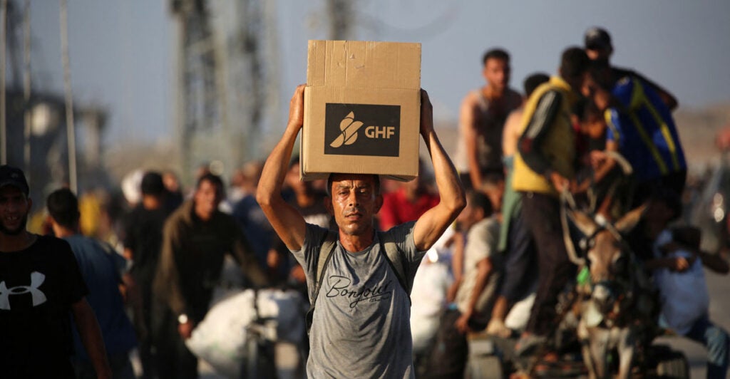 A man in a grey shirt carrying a Gaza Humanitarian Foundation aid parcel on his head walks along a road in the central Gaza Strip.