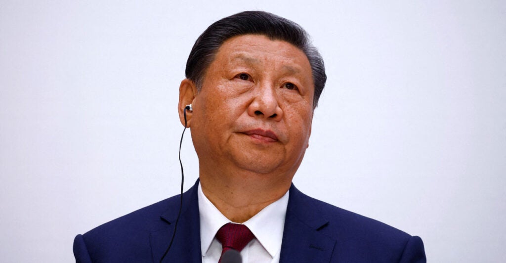 Xi Jinping looking up and to his left against a light gray background.