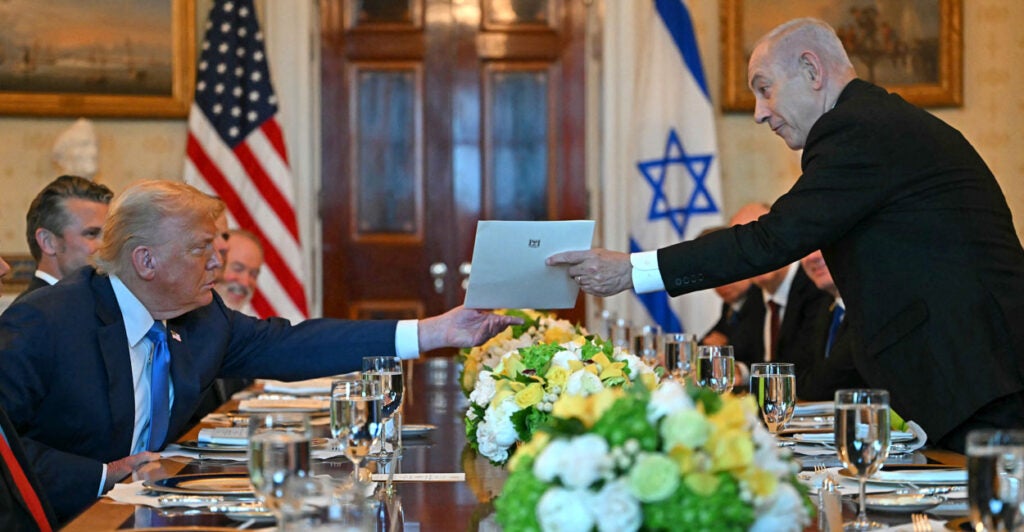 Benjamin Netanyahu stands at an elegant dinner table handing a letter to a seated President Trump, with an American and an Israeli flag in the background.