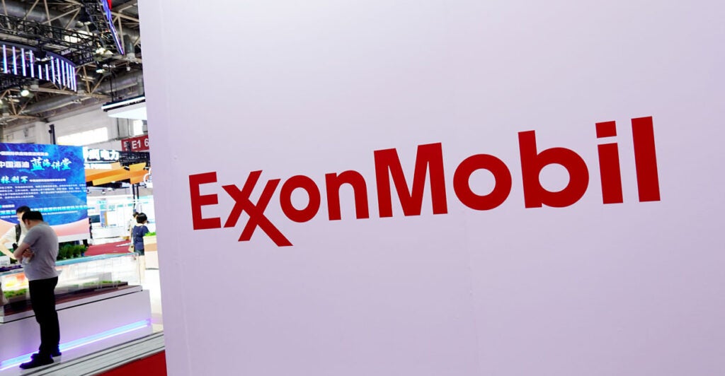 Close-up of an ExxonMobile logo on a white background, with exhibits and two attendees at 3rd China International Supply Chain Expo on the left side of the photo.