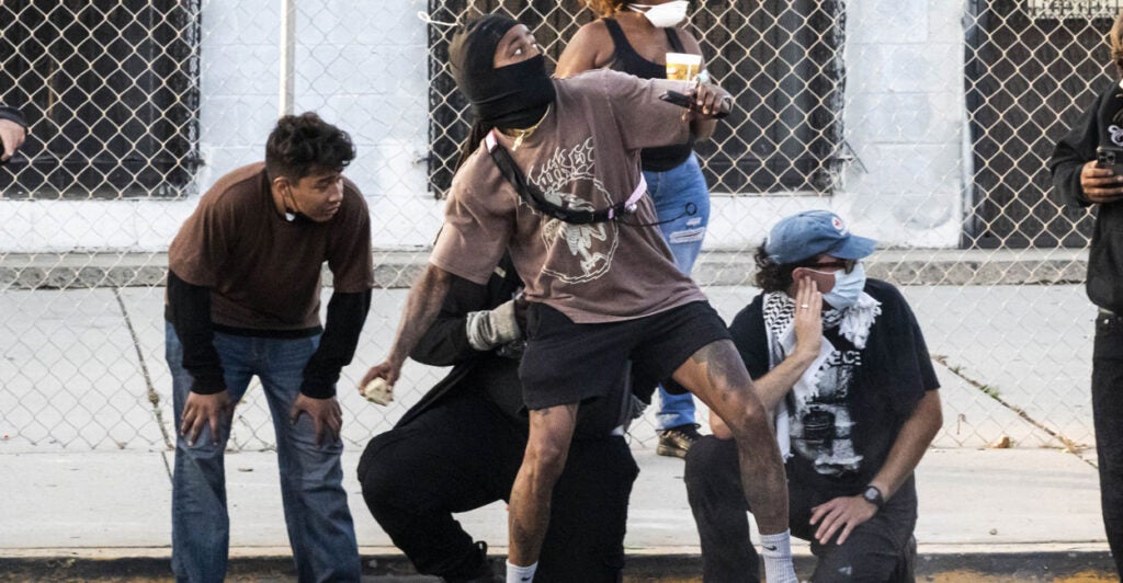 Tattooed ICE protester in mask throws object, as others look on.