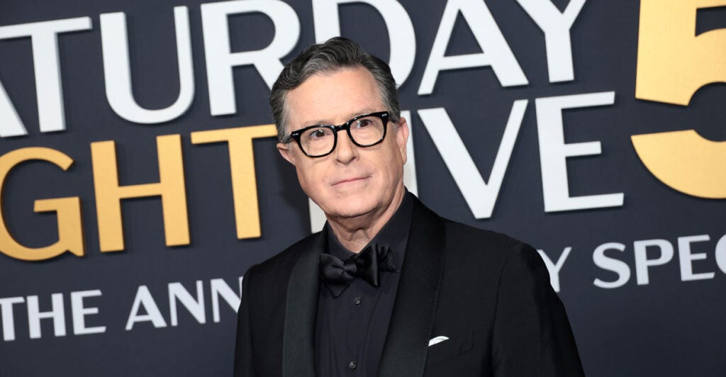 Stephen Colbert stands in a black suit with black shirt in front of "Saturday Night Live" 50th anniversary sign.