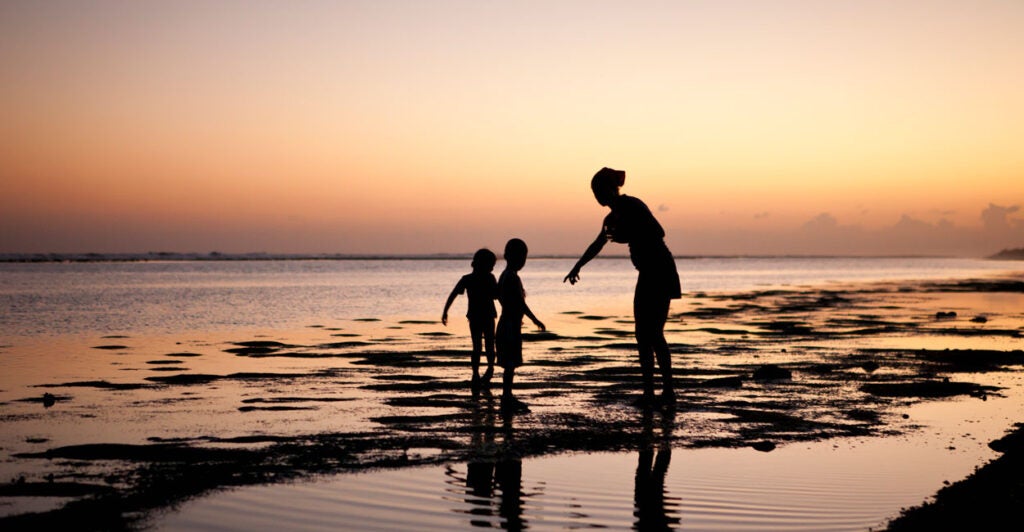 A mother and her two kids in silhouette at sunset their feet in the ocean.