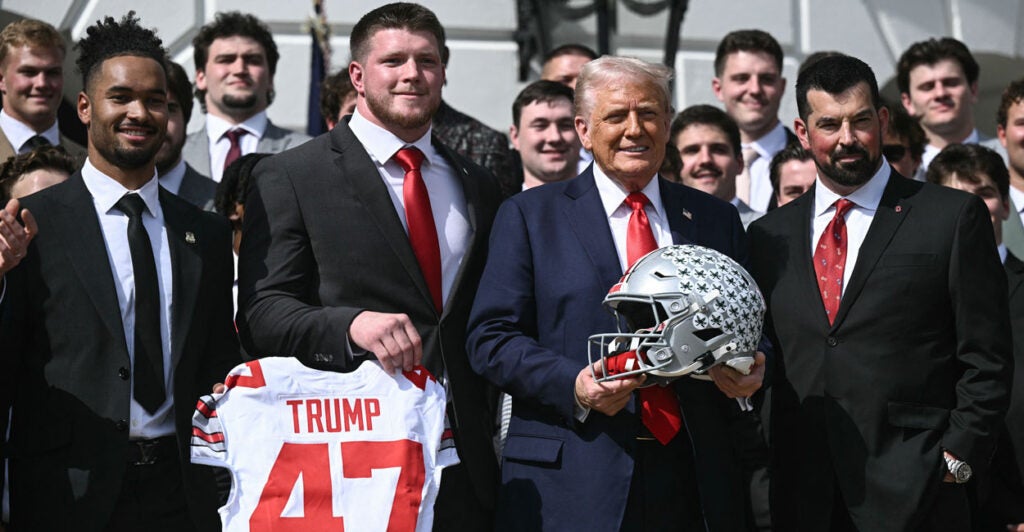 President Donald Trump holds an Ohio State football helmet while surrounded by the NCAA championship football team in front of the White House.