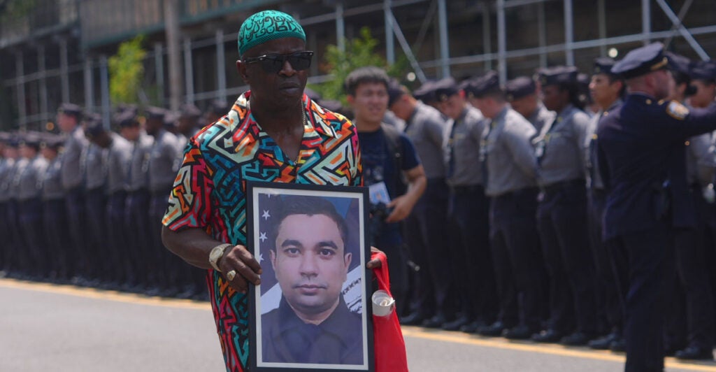 A man in colorful shirt holds a picture of slain NYPD officer Didarul Islam during his funeral procession. Along the street a line of officers bow their heads.