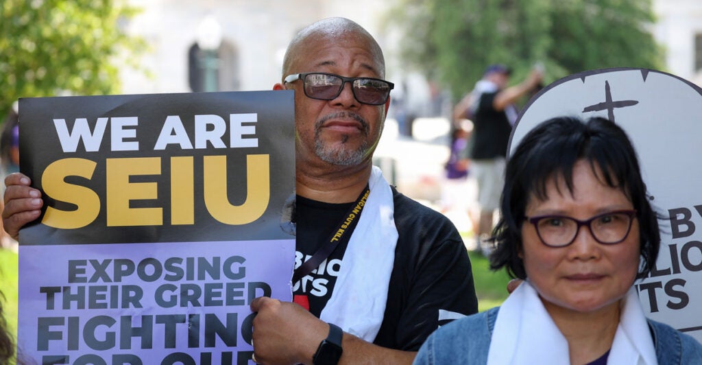 Male SEIU member holds sign "We Are SEIU: Exposing Their Greed" while standin g next to woman with towel around her neck.