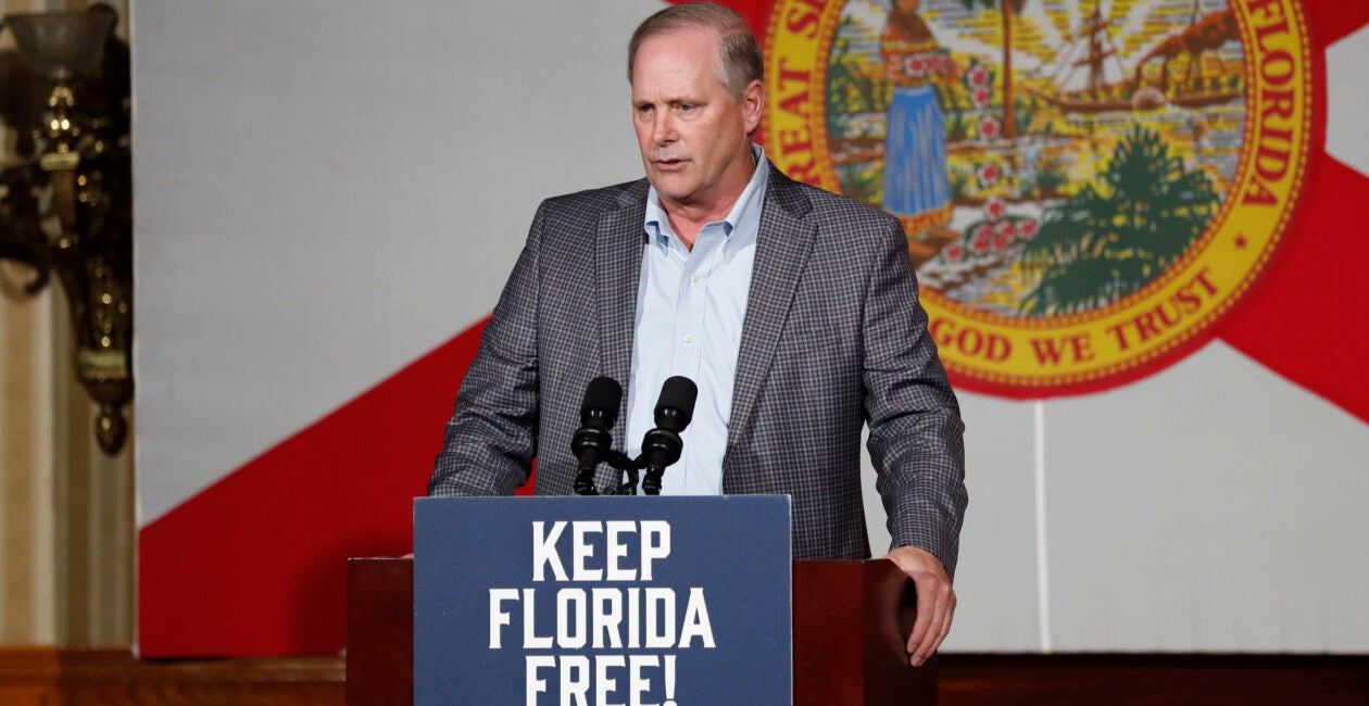 Florida Agriculture Commissioner Wilt Simpson in a suit jacket stands at a podium with the sign that says keep Florida free