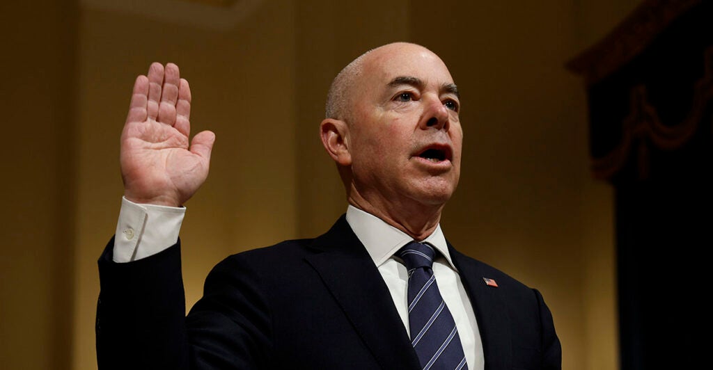 Alejandro Mayorkas in a black suit raising his hand.