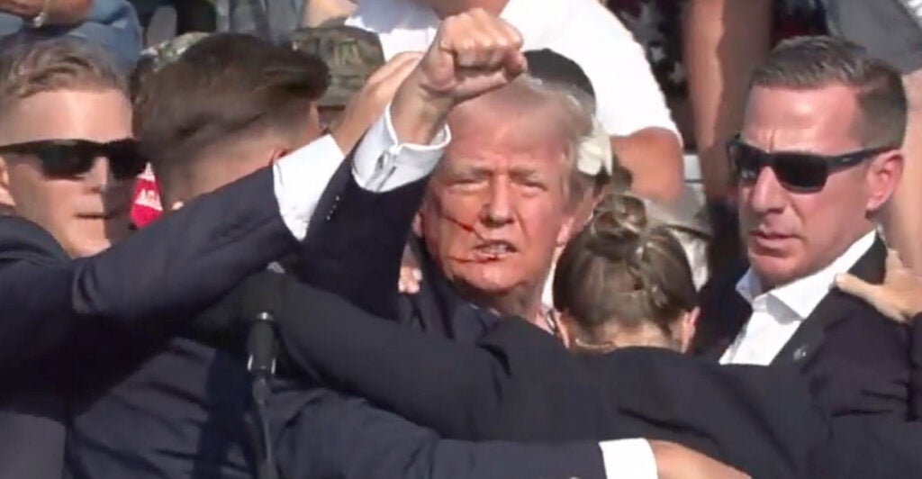 A blooded Donald Trump raises his fist while being assisted by Secret Service agents after being wounded in July 13, 2024 assassination attempt.