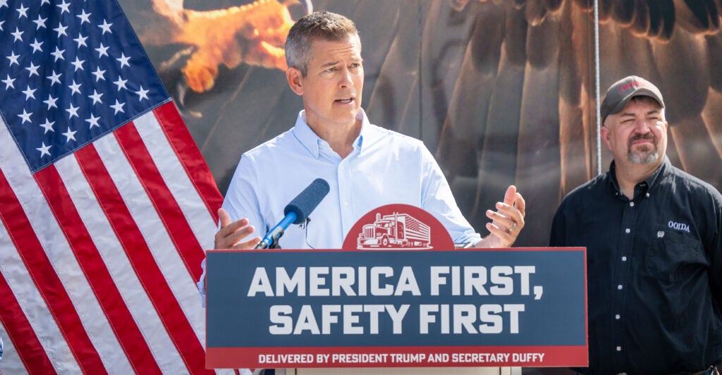 Transportation Secretary Sean Duffy In a collared shirt speaks Outdoors at a podium that has a sign that says America first, safety first