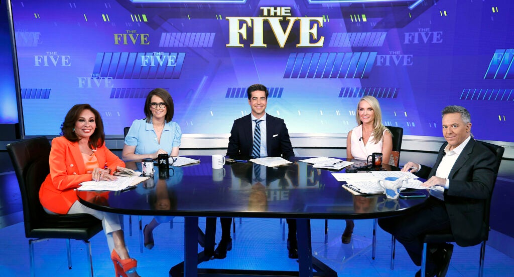 Greg Gutfeld, Dana Perino, Jesse Waters, Jessica Tarlov and Jeanine Pirro sit at the table on the set of "The Five" looking straight to the camera.