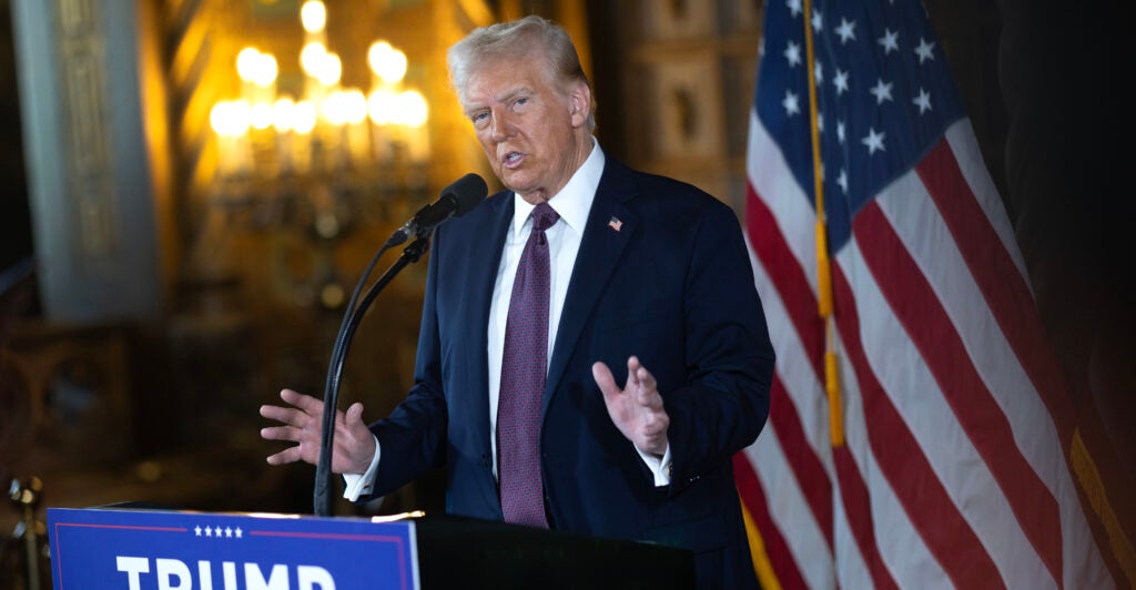 President Donald Trump in a dark suit speaks at a podium at his Mar-a-Lago Club. A US flag is in the background