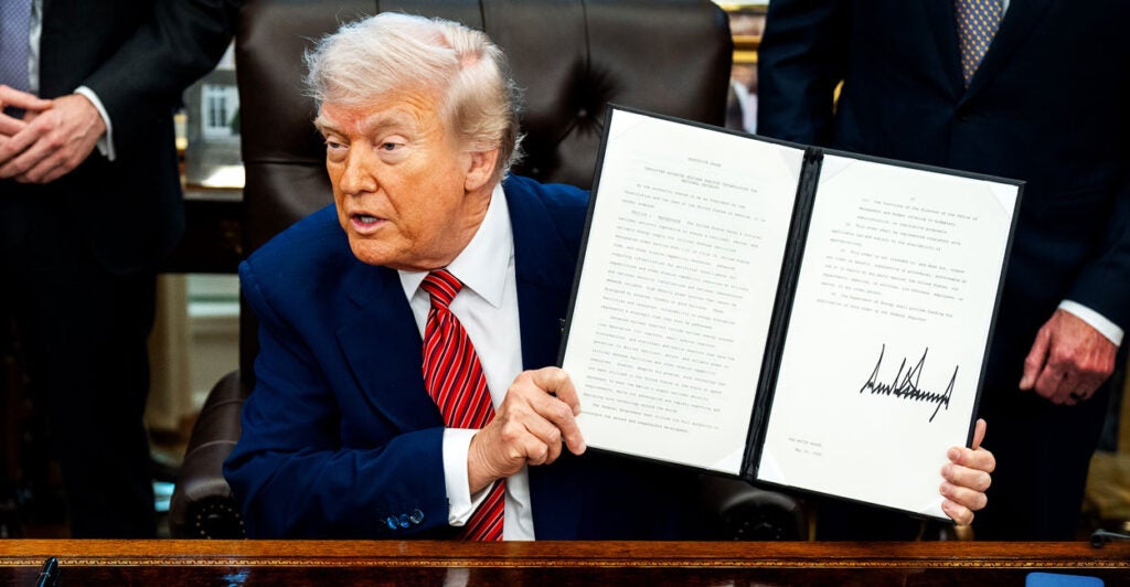 Donald Trump in a blue suit and red tie holds up an executive order he just signed in the Oval Office at his desk