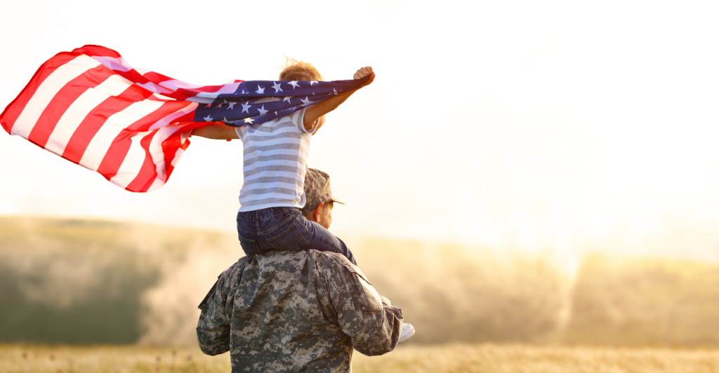 Rear view of military father carrying son holding American flag on his shoulders